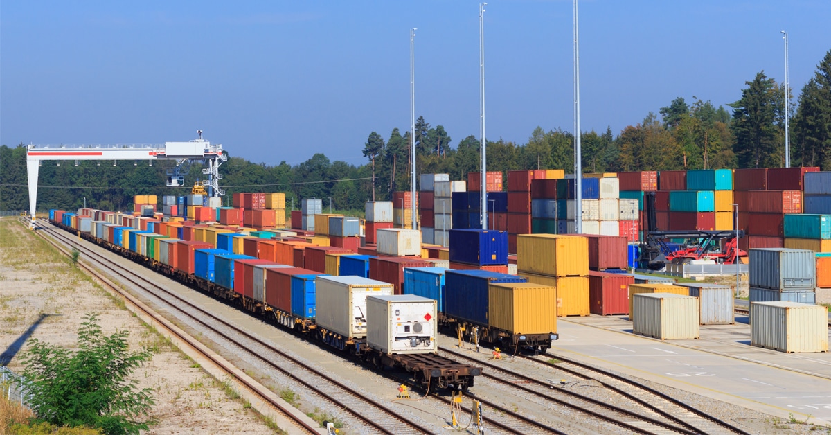 A series of colorful rail boxcars along a sandy rail yard with some being loaded by a crane onto a train.