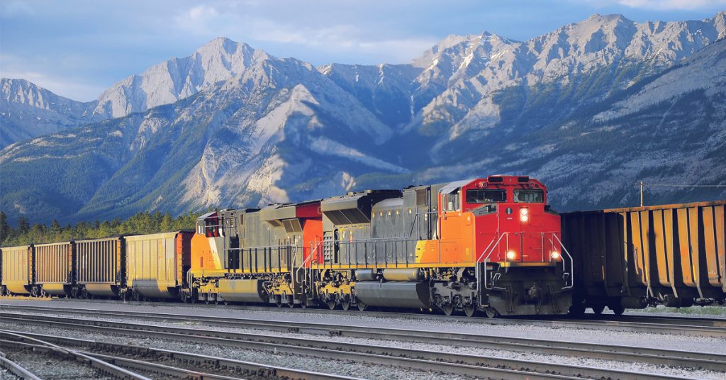 A red, yellow, and black rail engine hauling yellow cars on a track with snow covered mountains in the background.
