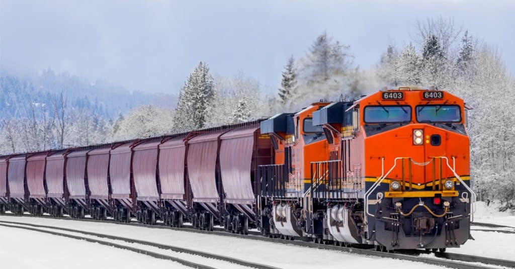 An orange rail freight engine hauls a line of brown rail cars through a snowy forest.
