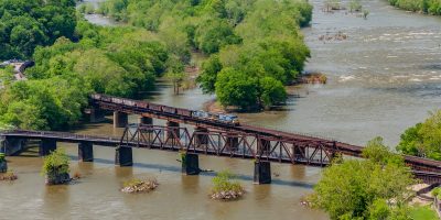 A blue rail freight engine with a line of rail cars crosses over a river on two rail infrastructure bridges.