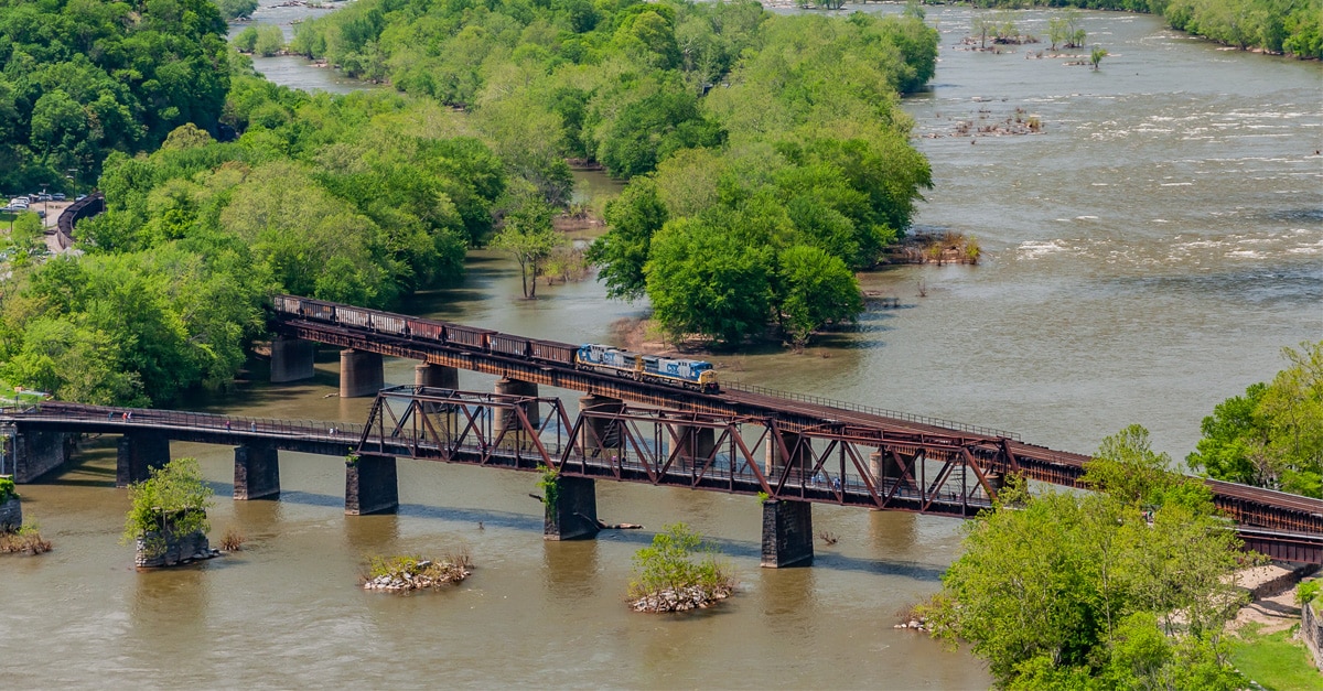 A blue rail freight engine with a line of rail cars crosses over a river on two rail infrastructure bridges.