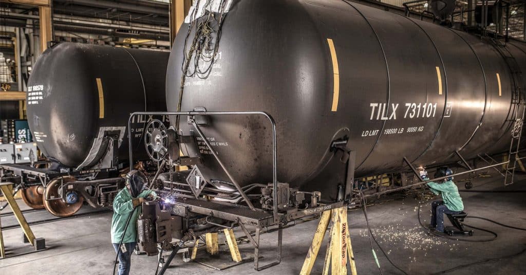 Two welders in green shirts work on a rail tank car with sparks flying.