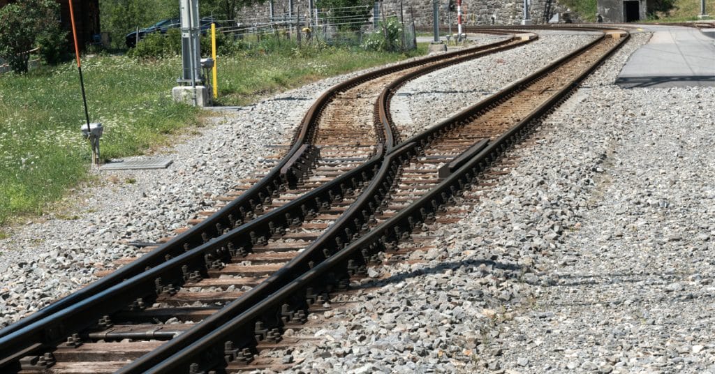 Two rail tracks over gravel and grass merges into one track.