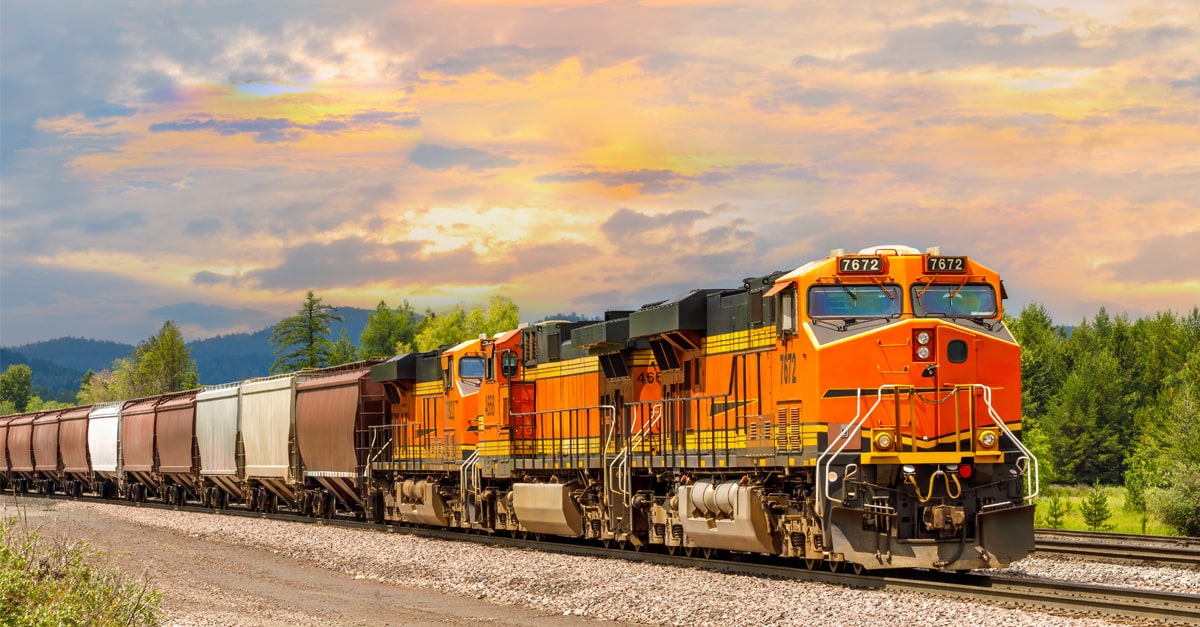 An orange rail engine hauls a line of brown and gray railcars through green trees.