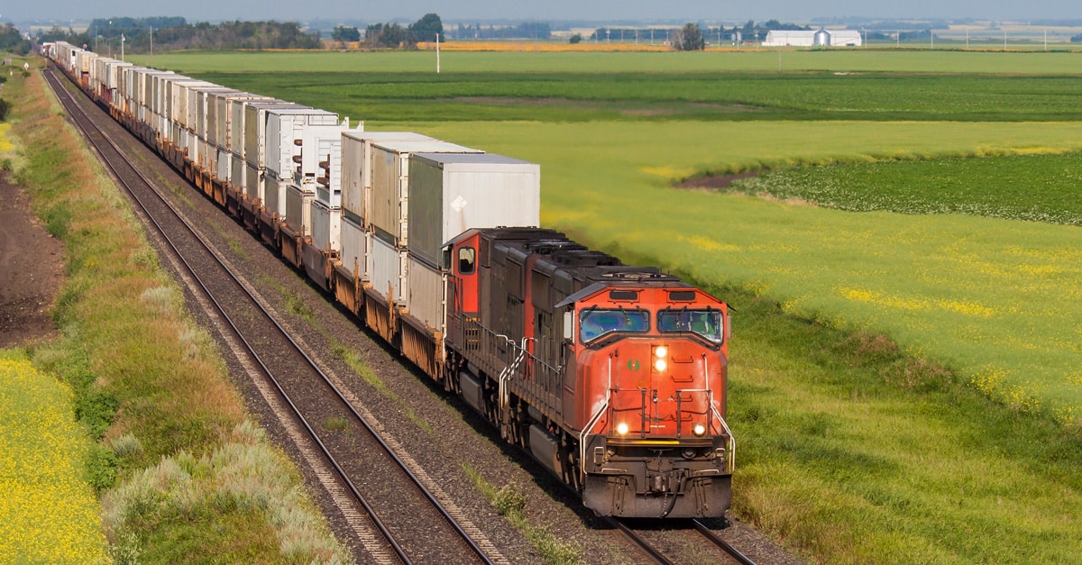 An orange locomotive hauls white rail freight cars through fields of green grass.