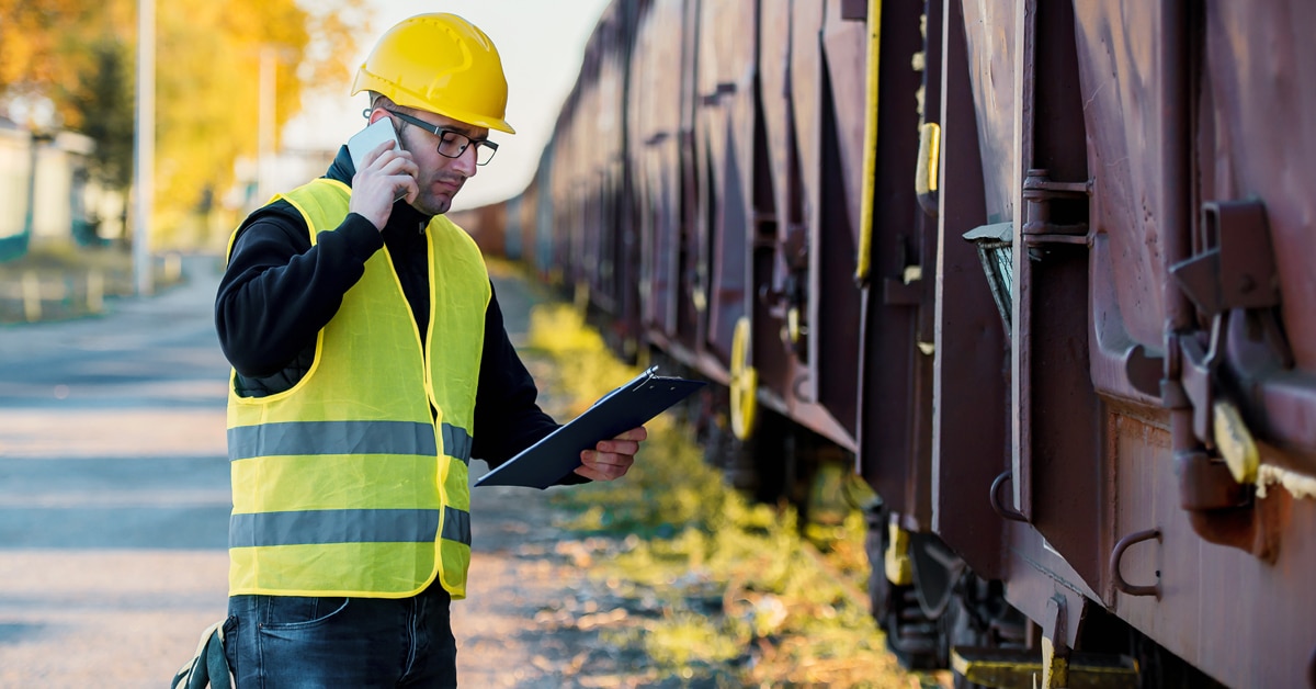 Rail worker in yellow safety vest speaking on phone and looking at tablet while beside freight boxcars.